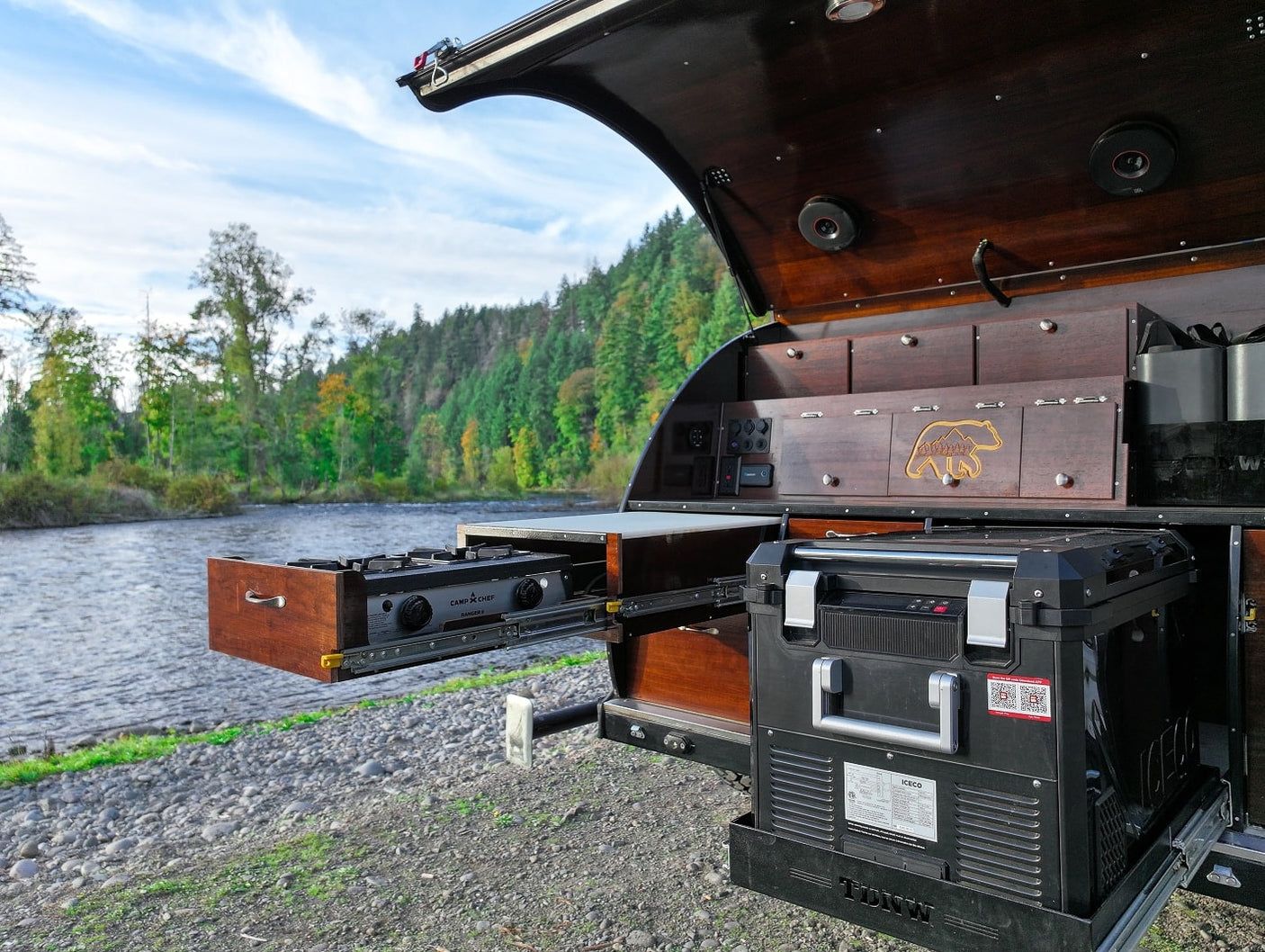 RV rental: A camper's open kitchen, featuring a pull-out stove and fridge, set against a beautiful river and forest backdrop.