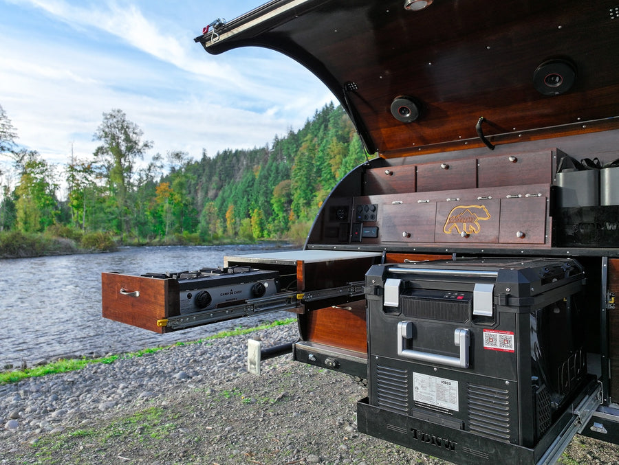 RV rental: A camper's open kitchen, featuring a pull-out stove and fridge, set against a beautiful river and forest backdrop.