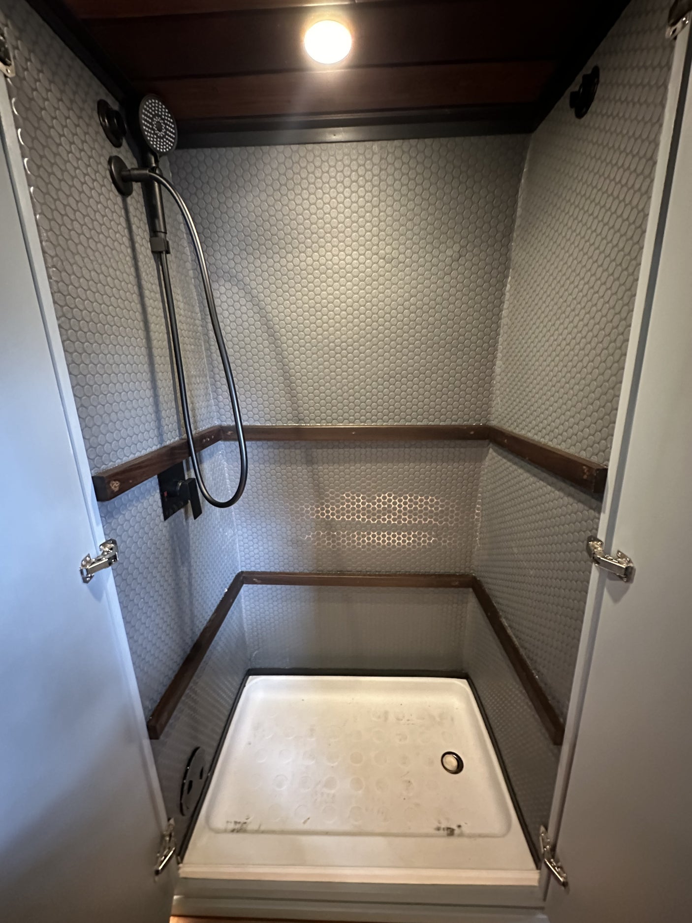 Compact RV rental shower stall with grey hexagon tiles, wood trim, and a white shower pan, featuring a black handheld shower.