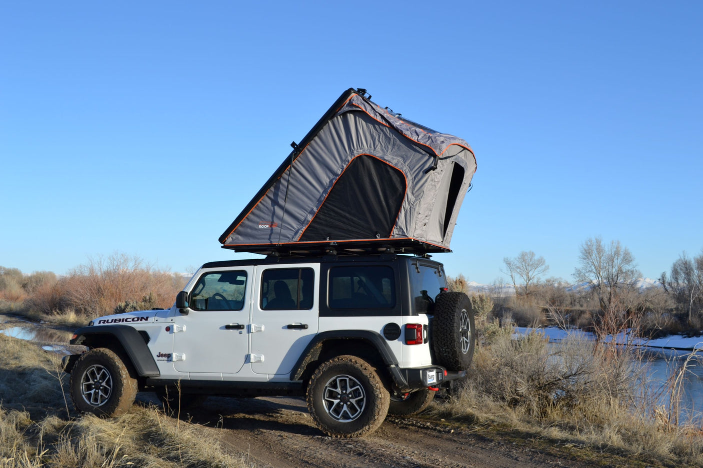 White Jeep Rubicon with grey rooftop tent deployed by a scenic winter river, perfect for an adventurous RV rental.