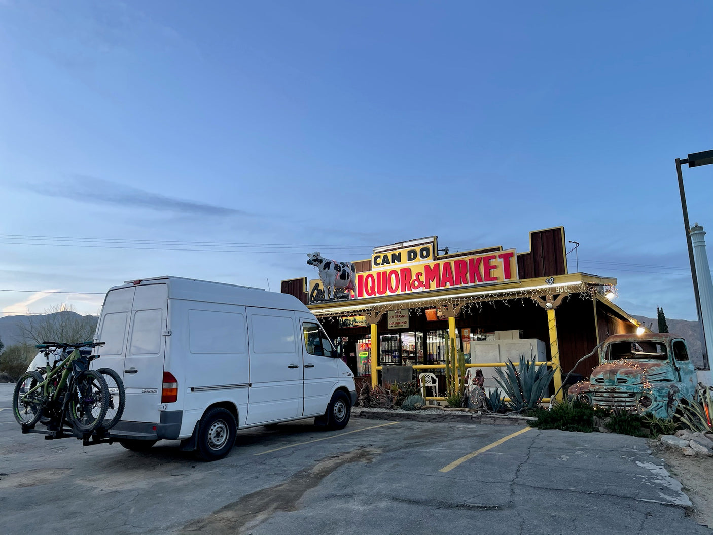 White Sprinter van, an RV rental option, carrying bikes, parked at Can Do Liquor & Market in a desert setting.