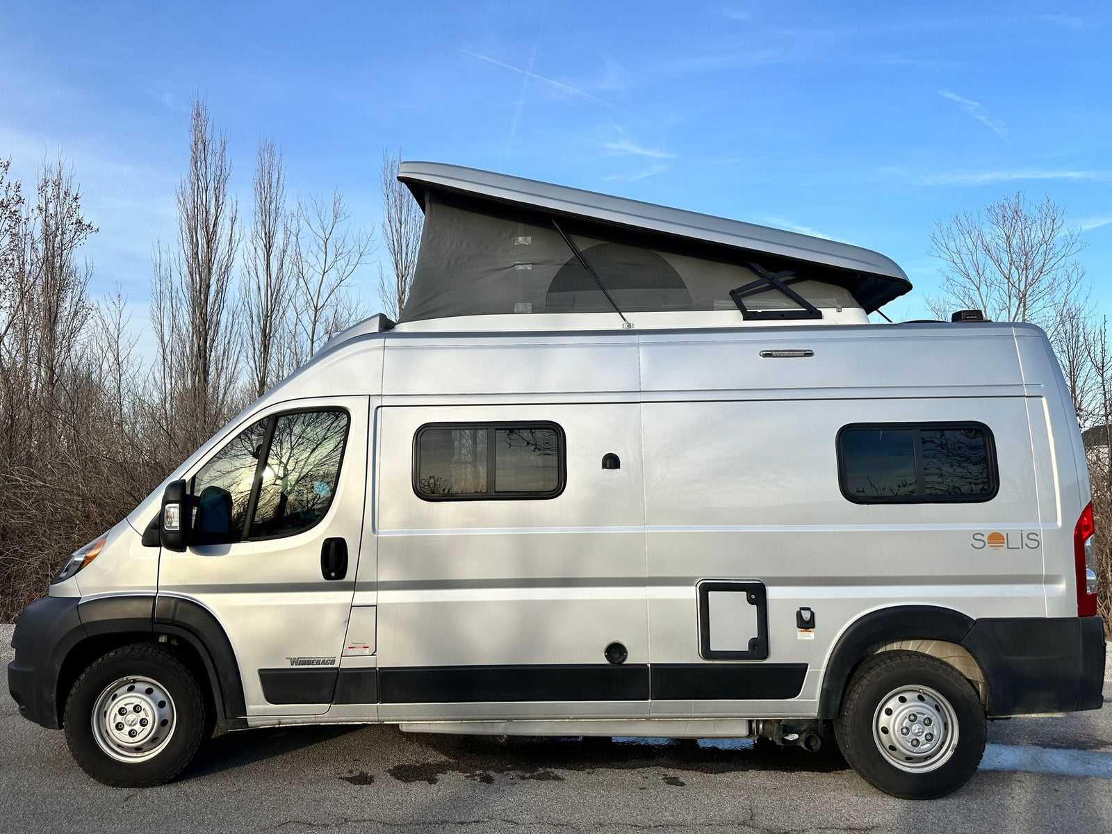 Silver Winnebago Solis camper van with pop-top for rv rental. Parked outdoors with bare trees & blue sky.|Sleek silver Winnebago Solis pop-top camper van parked