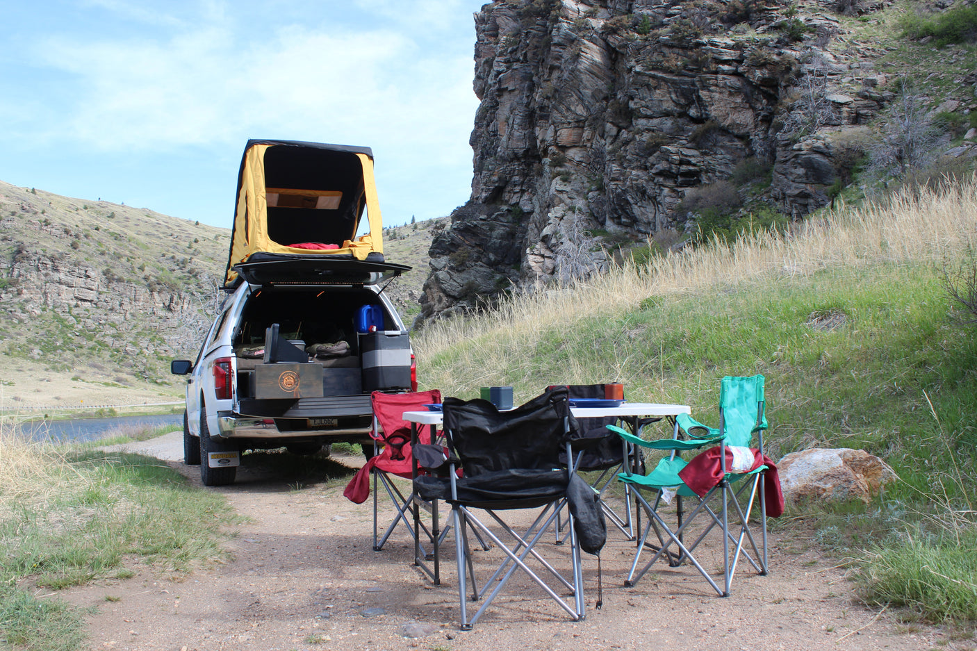 A truck with a rooftop tent, camping chairs, and table set up by a scenic lake. An ideal outdoor adventure for any RV rental!
