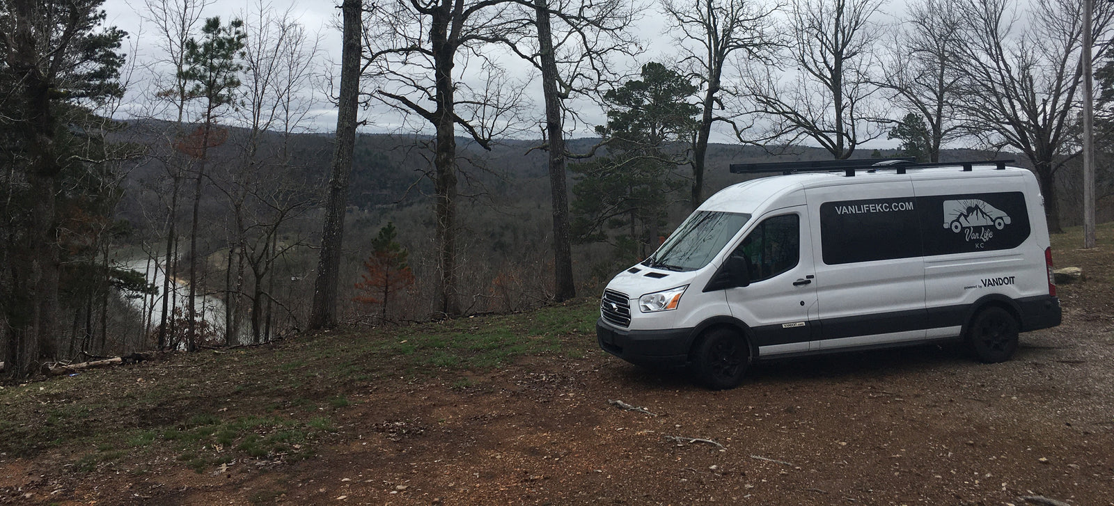 White Van Life KC RV rental on a dirt path overlooking a scenic wooded valley and lake.
