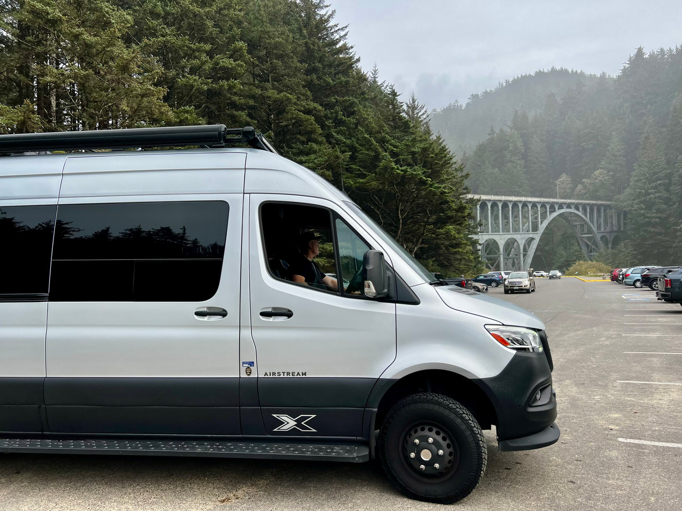 An Airstream RV rental parked at a scenic viewpoint, with a grand arched bridge and foggy evergreen forest in the background.