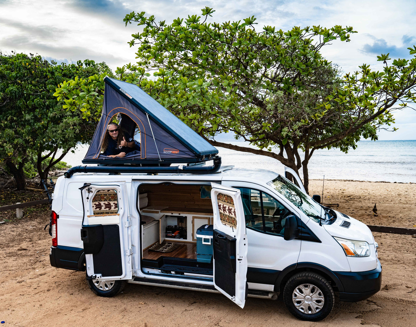 Experience an **rv rental**! Man in rooftop tent atop a white camper van, open doors reveal interior, parked by a sandy beach.