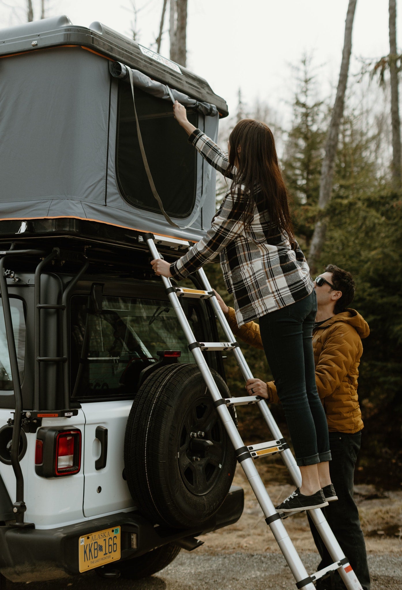A woman on a ladder adjusts a rooftop tent on her RV rental Jeep, with a man assisting. Perfect for your next camping adventure.