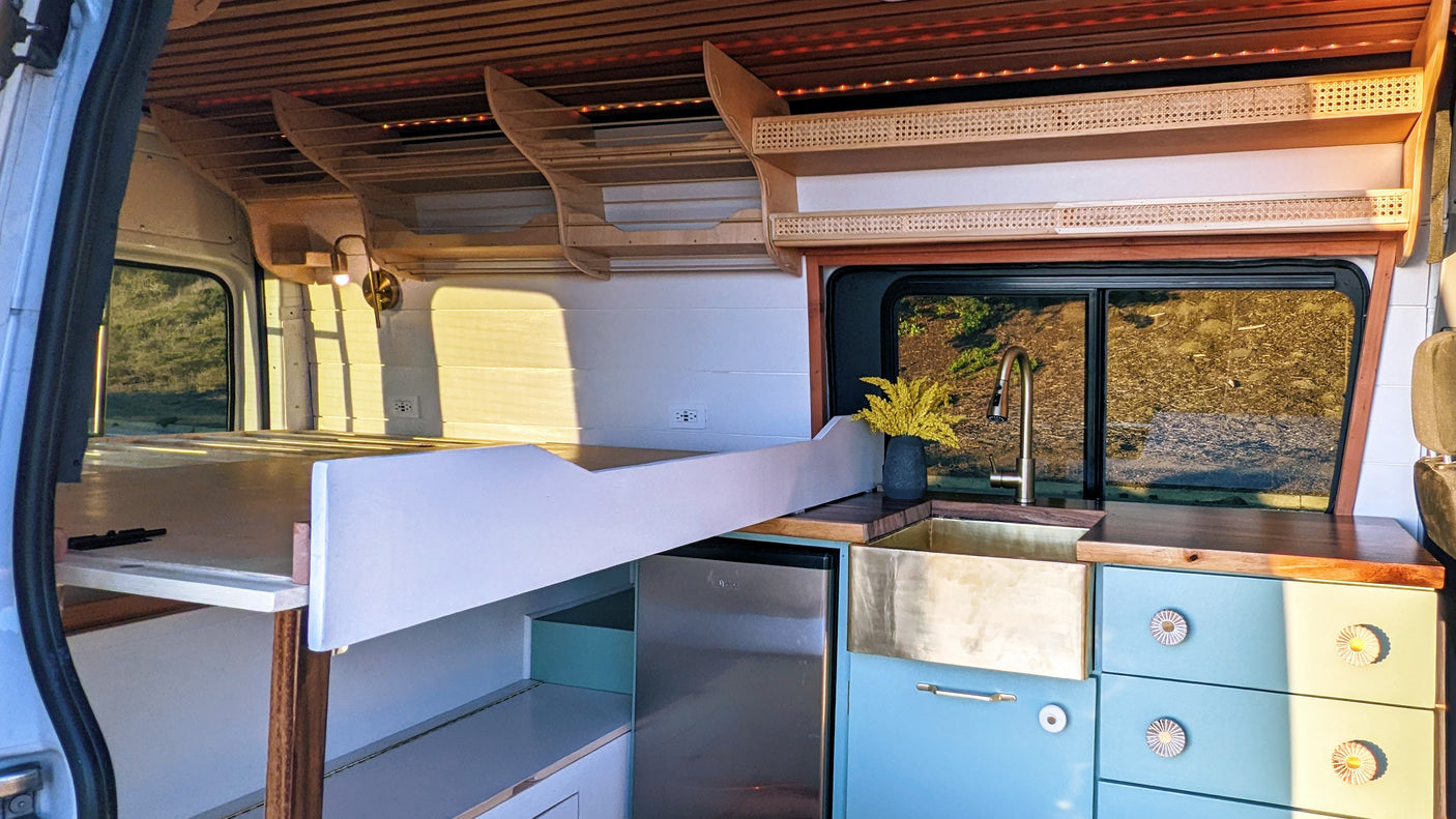 Inside a cozy RV rental: a modern kitchen with a brass sink, light blue cabinets, a comfortable bed, and custom wood shelving.