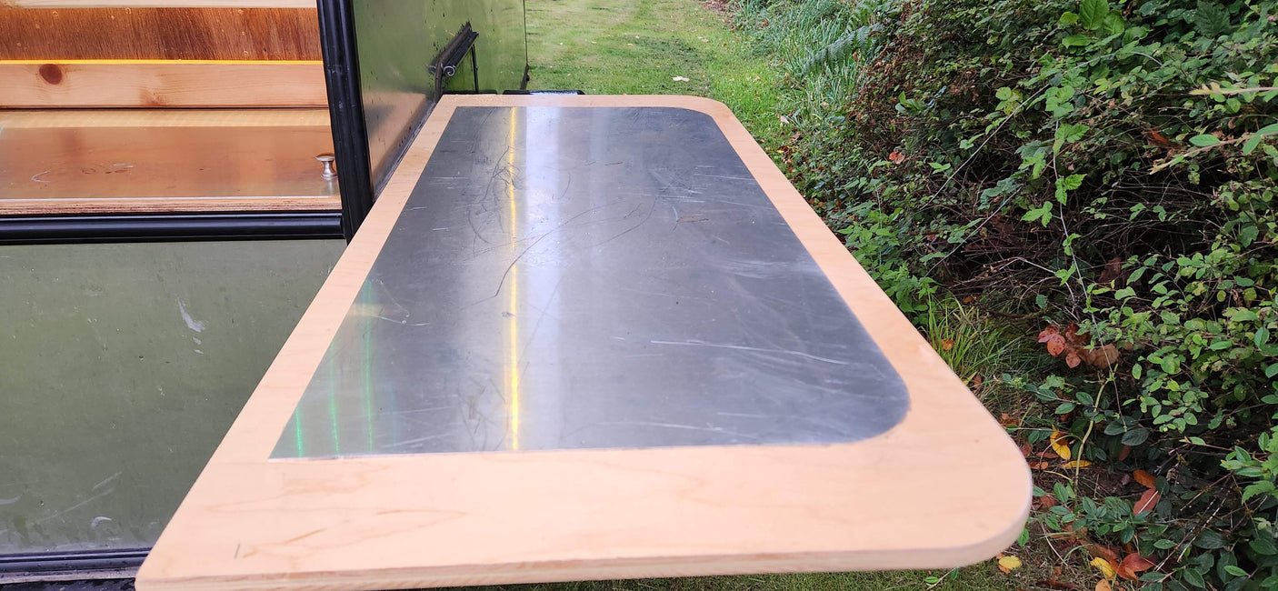 RV rental: Extendable outdoor table with scratched metal top & wood trim, against green foliage.