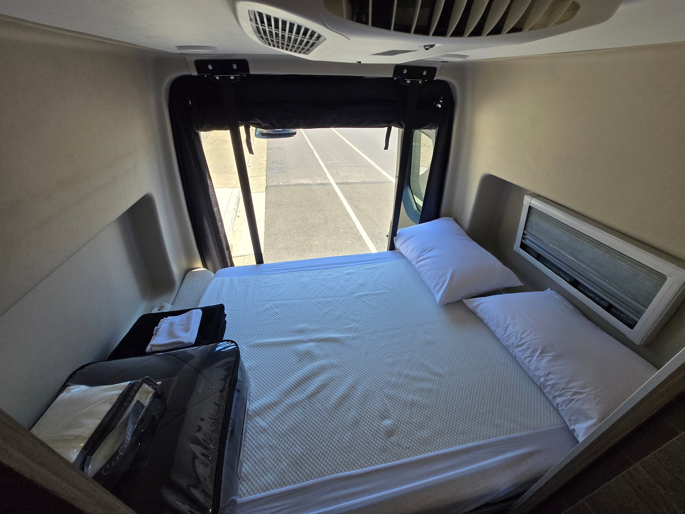 RV rental bedroom showcasing a bed with white pillows, a window view of the road, and an overhead AC unit.