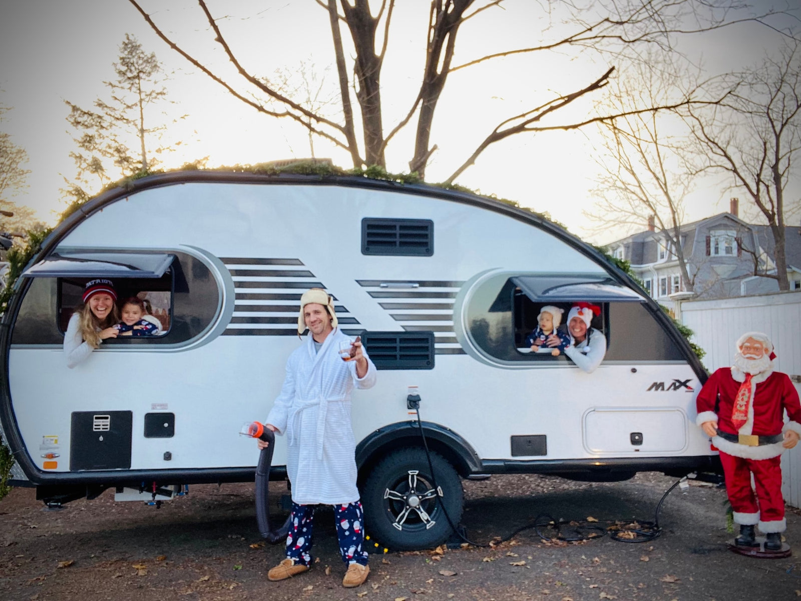 Joyful family holiday scene with an **RV rental**. Dad in PJs, mom & kids peeking from the festive teardrop camper windows, Santa nearby.