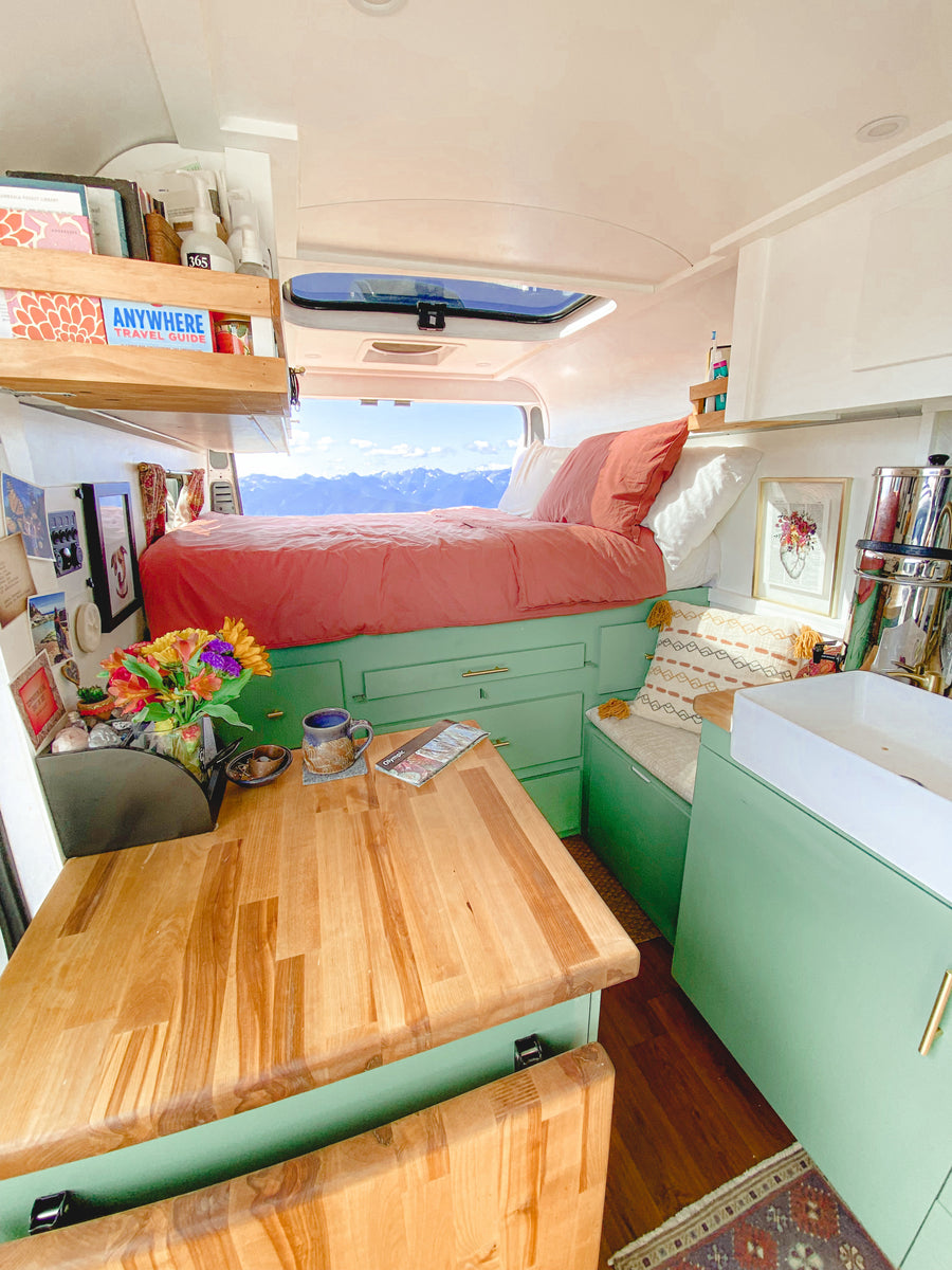 Cozy RV rental interior with a bed facing a stunning mountain view. Features sage green cabinets, a wooden table, and small kitchen.