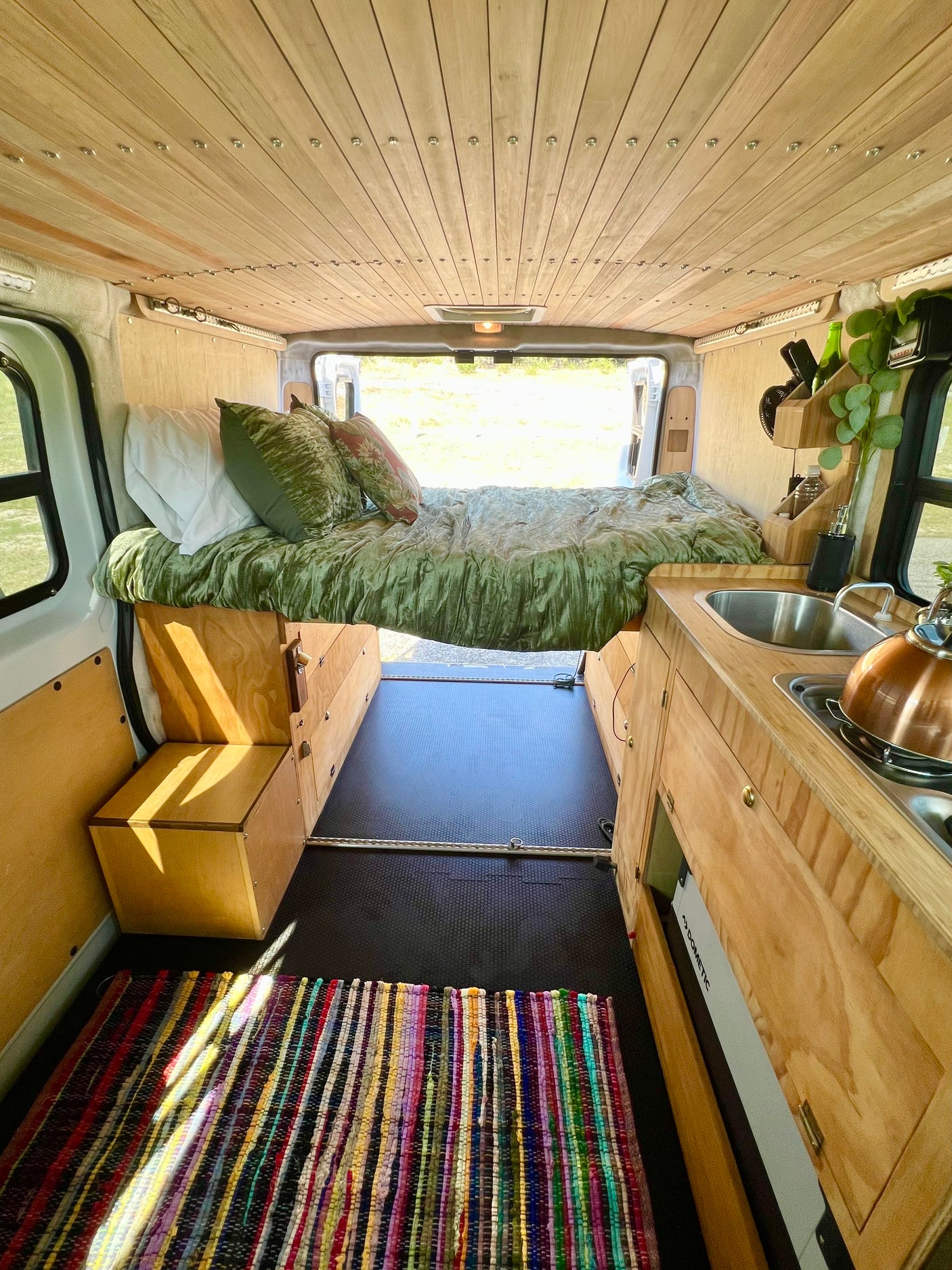 Inside a stylish **rv rental**: custom wood interior, cozy bed with green bedding, and a compact kitchen with sink and kettle.