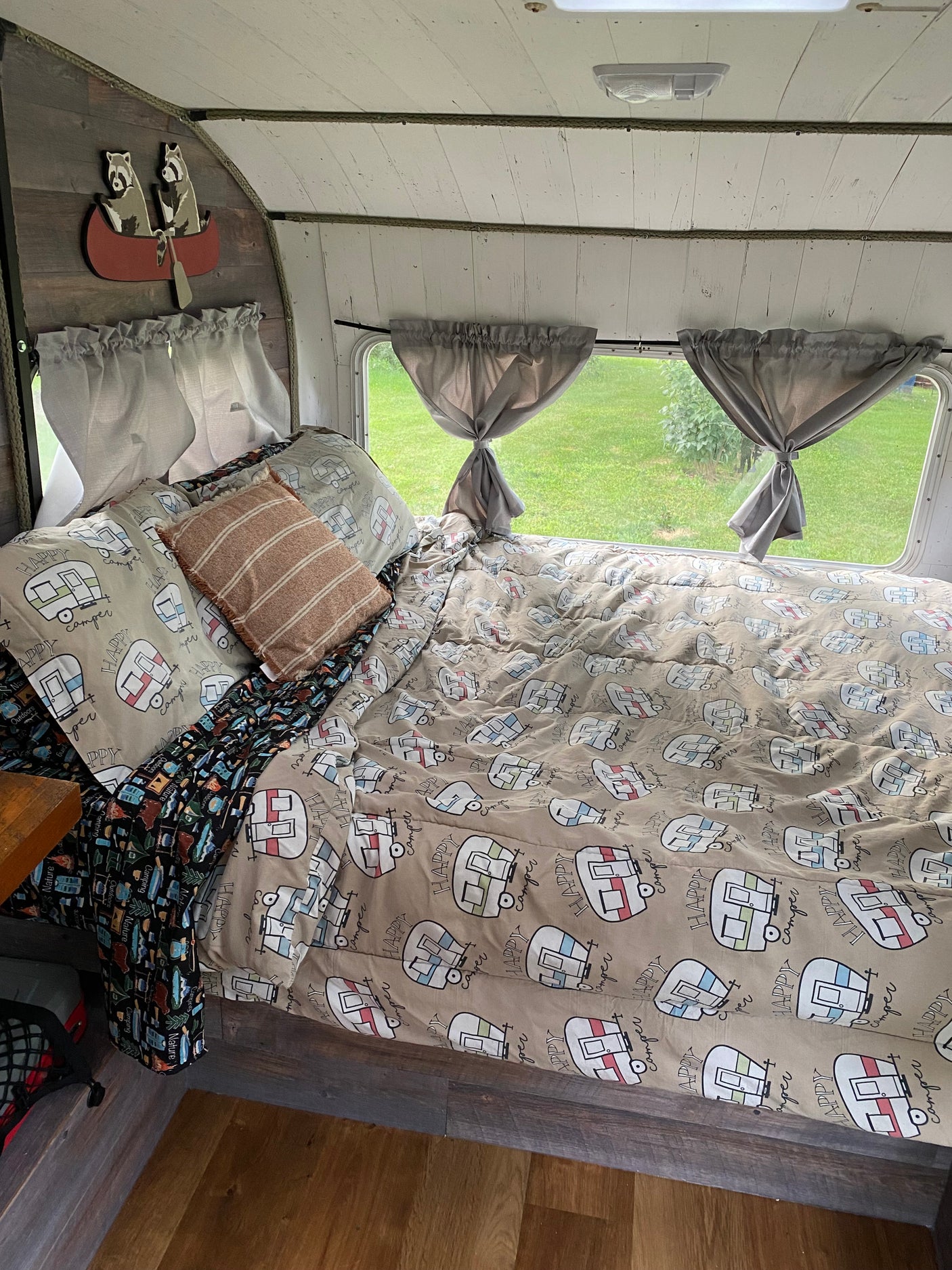 A cozy RV rental interior: "Happy Camper" bedding, charming raccoons in a canoe, and tied-back curtains.