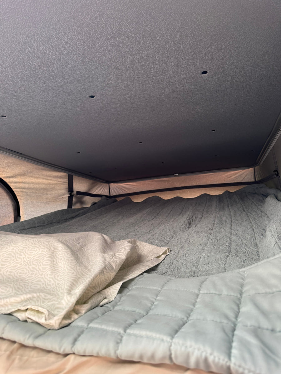 Inside an RV rental: a cozy bed with a patterned pillow, quilted blanket, and textured bedding under a ceiling with small lights.