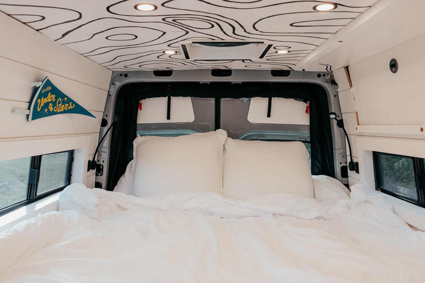 Cozy rv rental bedroom with custom topographic ceiling, plush white bed, and "Sleep Under the Stars" pennant.