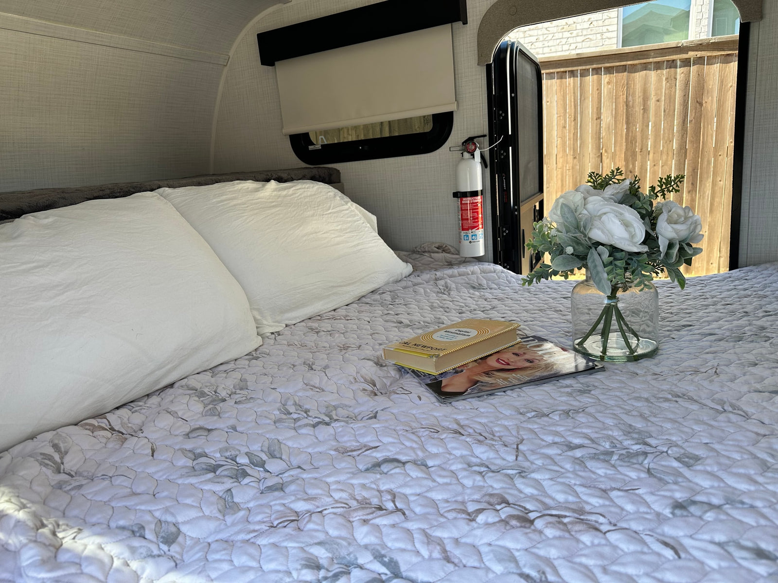 Cozy rv rental bedroom with a patterned quilt, white pillows, book, magazine, and flowers. Fire extinguisher visible near open door.