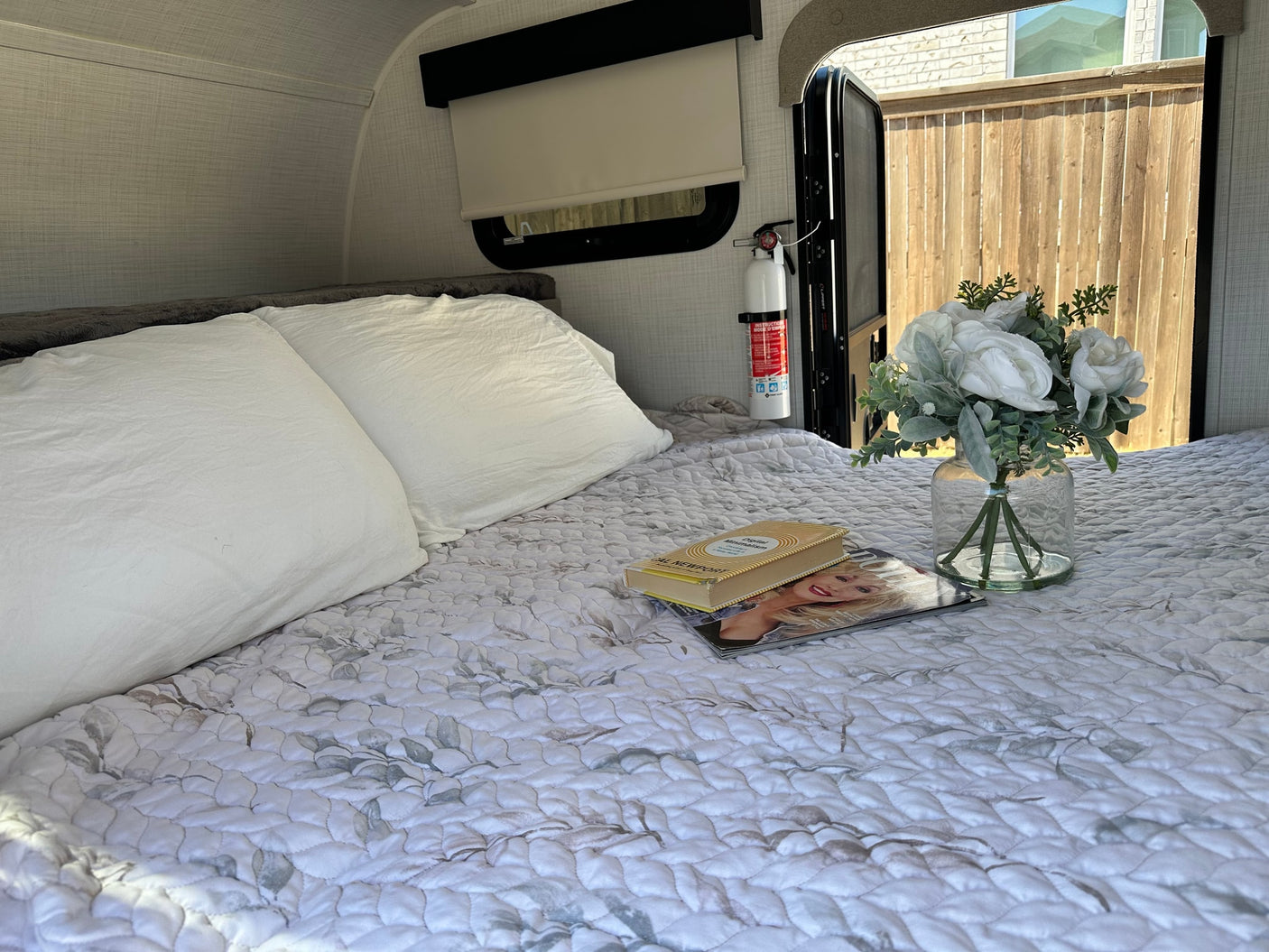 Cozy rv rental bedroom with a patterned quilt, white pillows, book, magazine, and flowers. Fire extinguisher visible near open door.