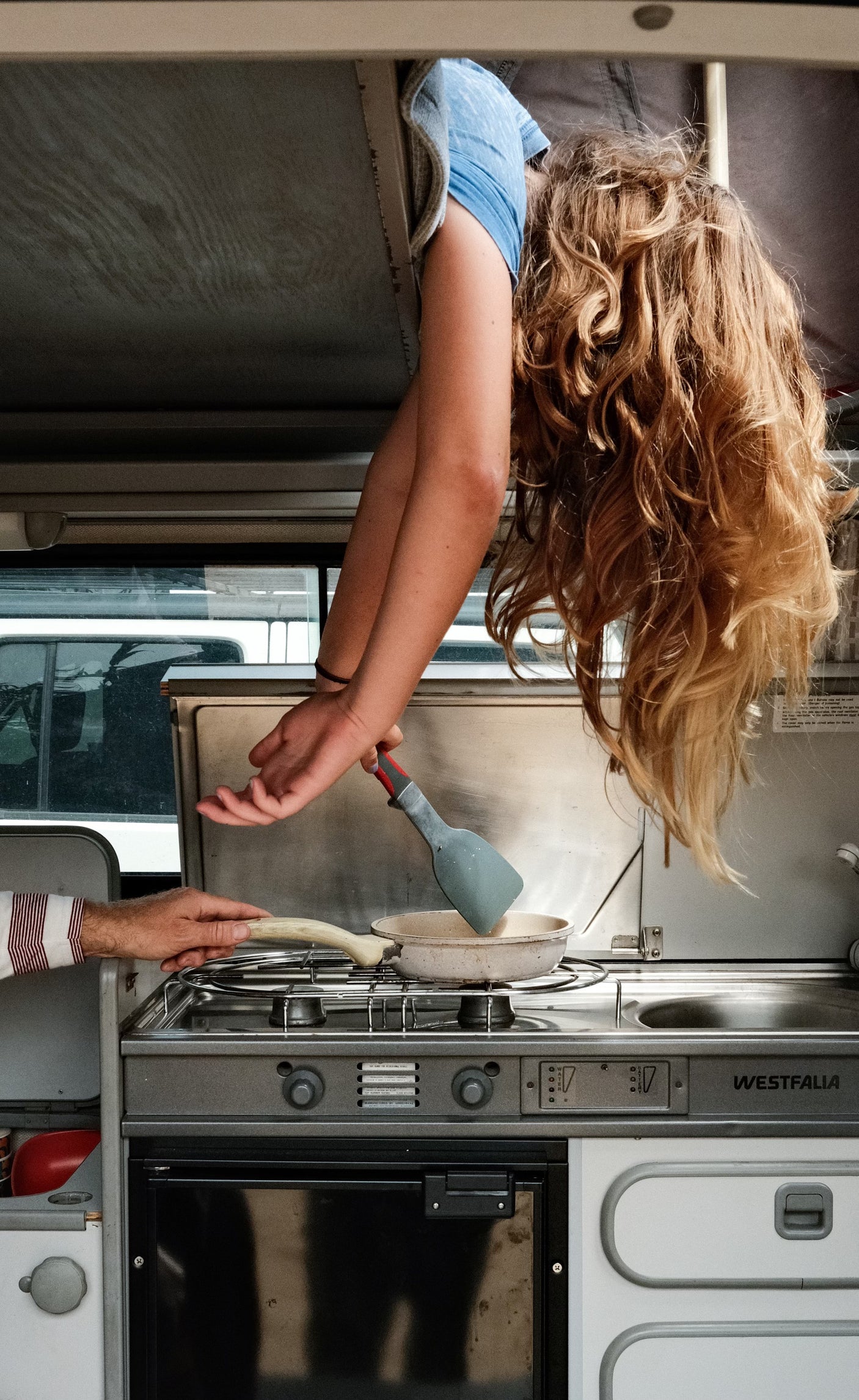Cooking in an RV rental: A person hangs upside down, long hair flowing, holding a spatula over a pan on a Westfalia stove.