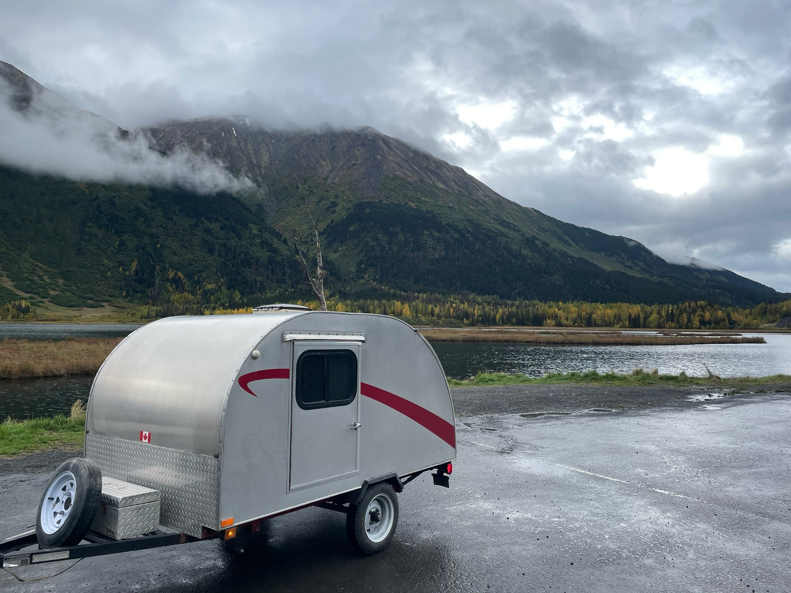 offering a picturesque nature escape.|Experience nature with this rv rental: a silver teardrop camper by a serene lake and misty mountains under cloudy skies.|Explore misty autumn mountains with this silver teardrop camper. An ideal rv rental for a cozy fall getaway amidst golden trees.|White SUV towing a teardrop rv rental camper by a misty lake and mountains under a cloudy sky