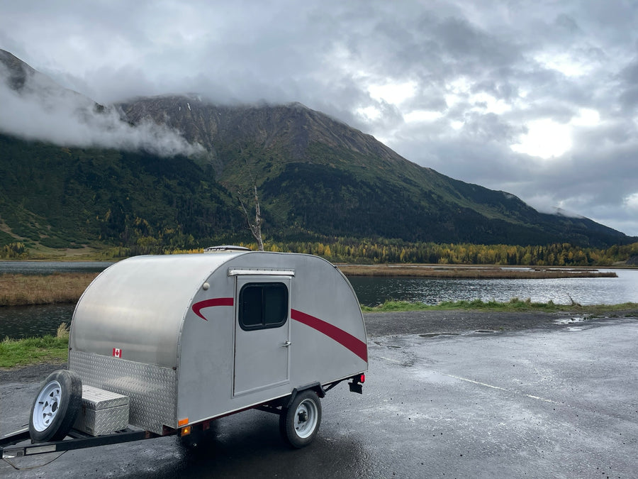 offering a picturesque nature escape.|Experience nature with this rv rental: a silver teardrop camper by a serene lake and misty mountains under cloudy skies.|Explore misty autumn mountains with this silver teardrop camper. An ideal rv rental for a cozy fall getaway amidst golden trees.|White SUV towing a teardrop rv rental camper by a misty lake and mountains under a cloudy sky