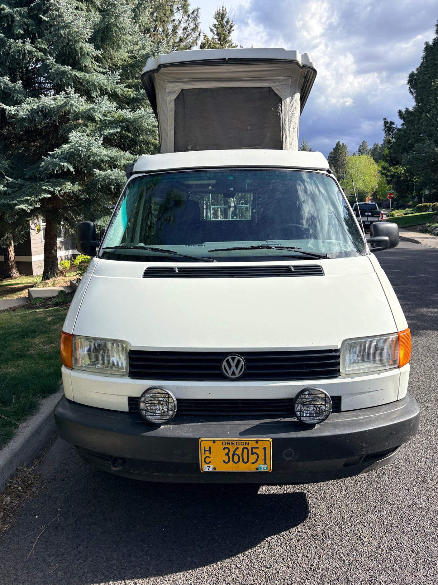 Front view of a white Volkswagen pop-top camper van, perhaps an RV rental, parked on a street by trees. Oregon plate visible.