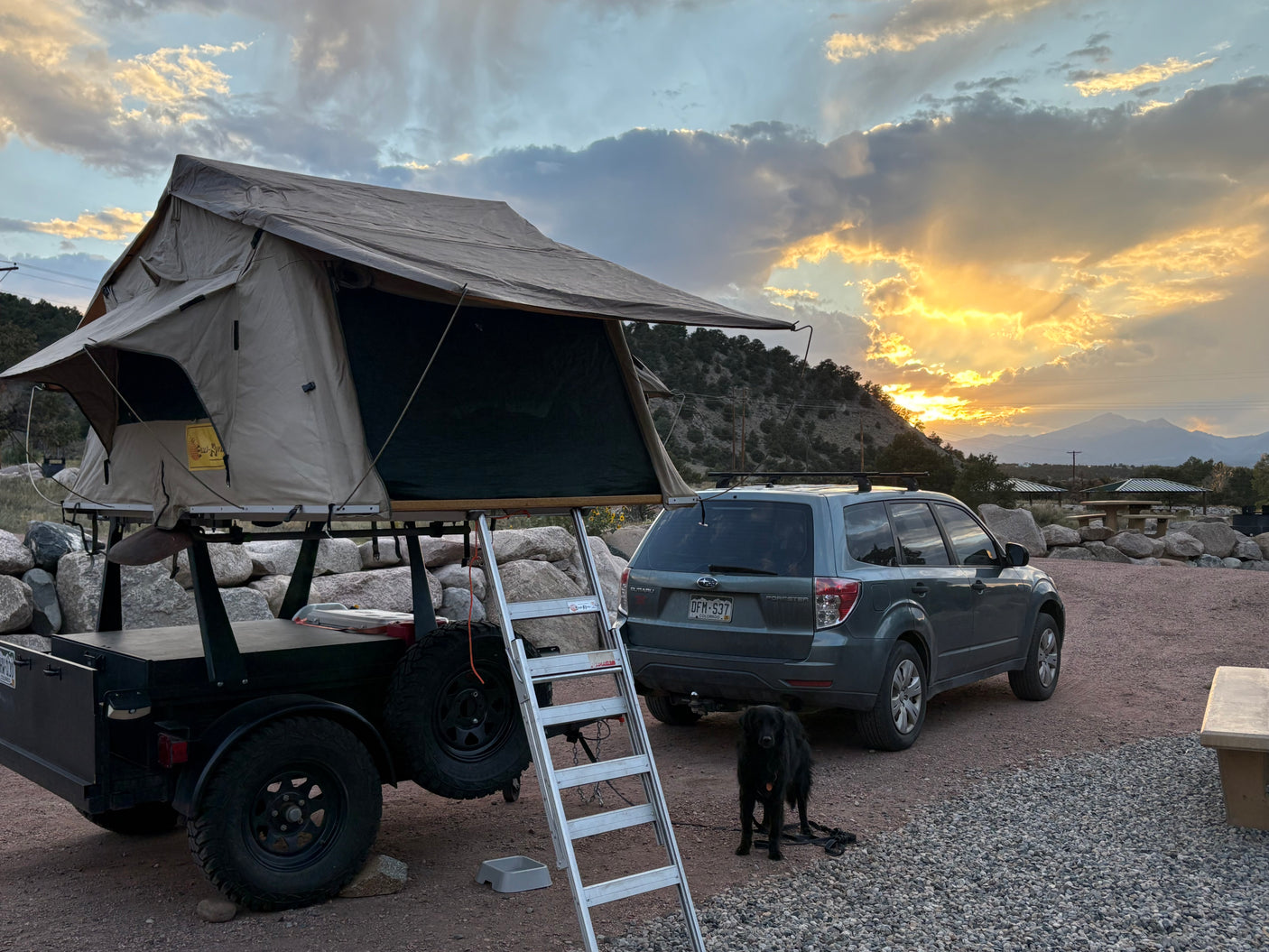 This RV rental adventure includes a rooftop tent, Subaru, and black dog, enjoying sunset at a mountain campsite.