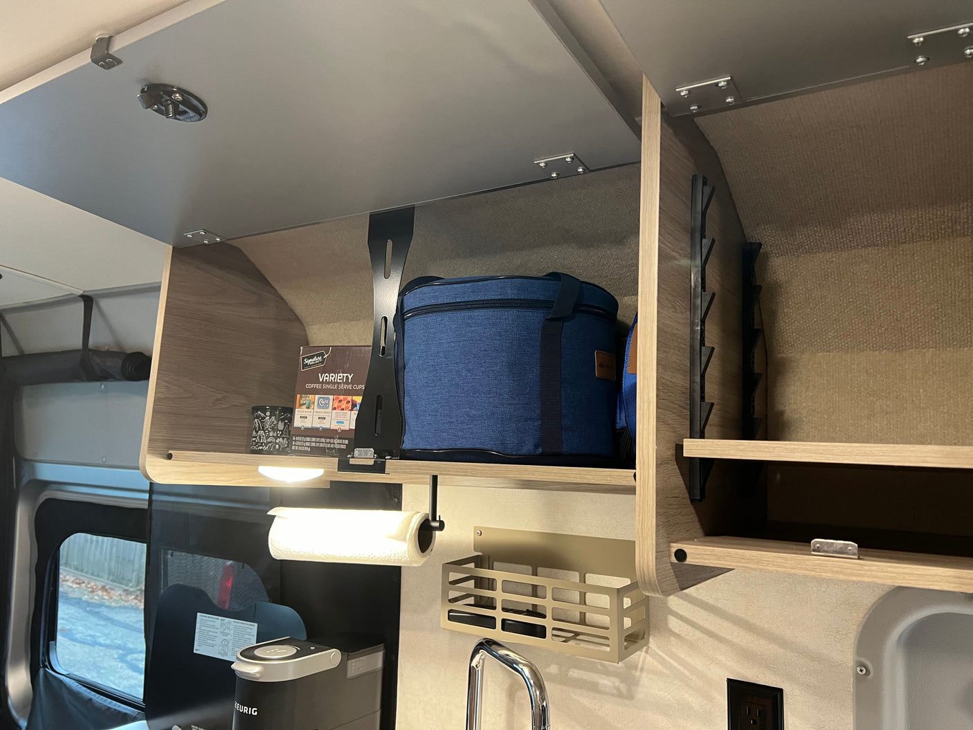 RV rental kitchen with open overhead cabinets showing coffee pods, a blue cooler bag, and a paper towel holder below a light.