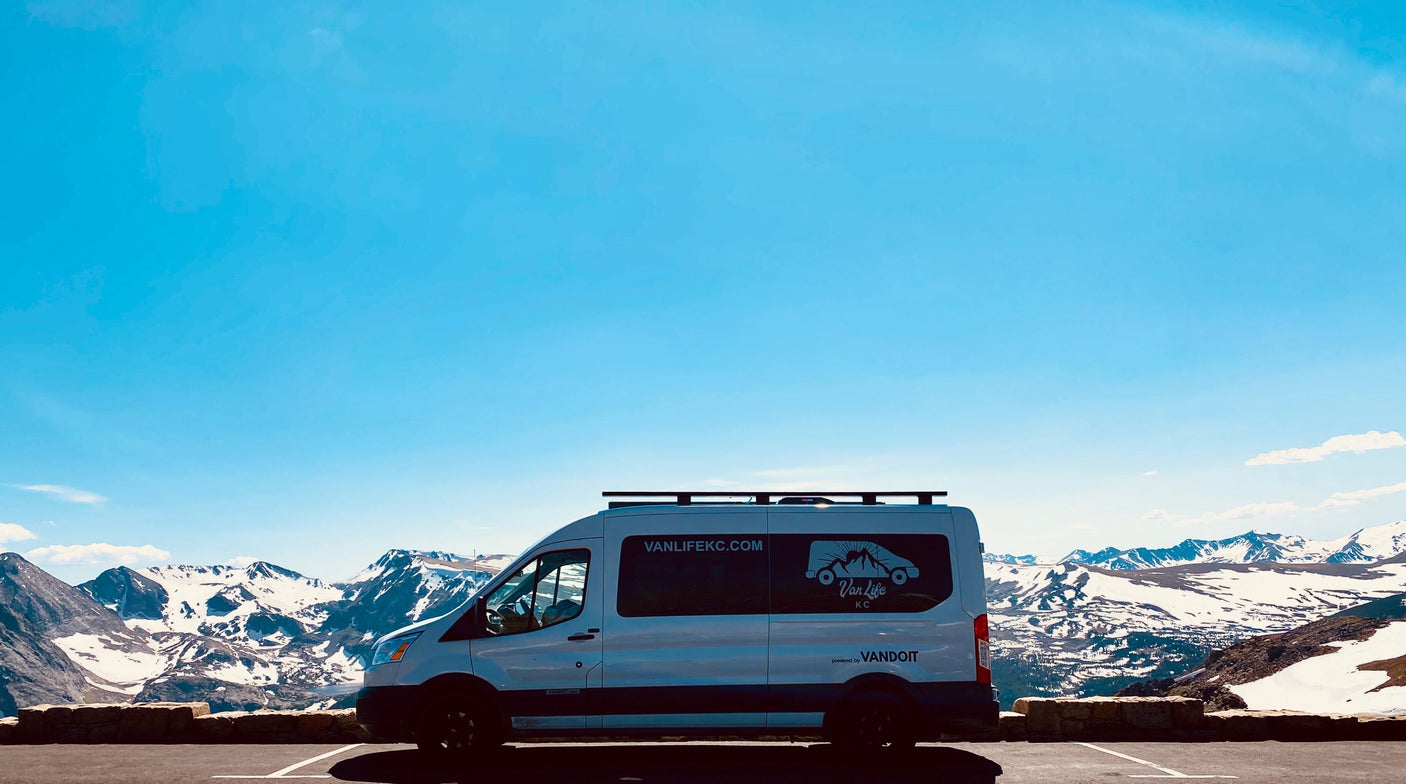 This white RV rental camper van, branded for Van Life KC, rests before grand snow-capped mountains under a clear blue sky.