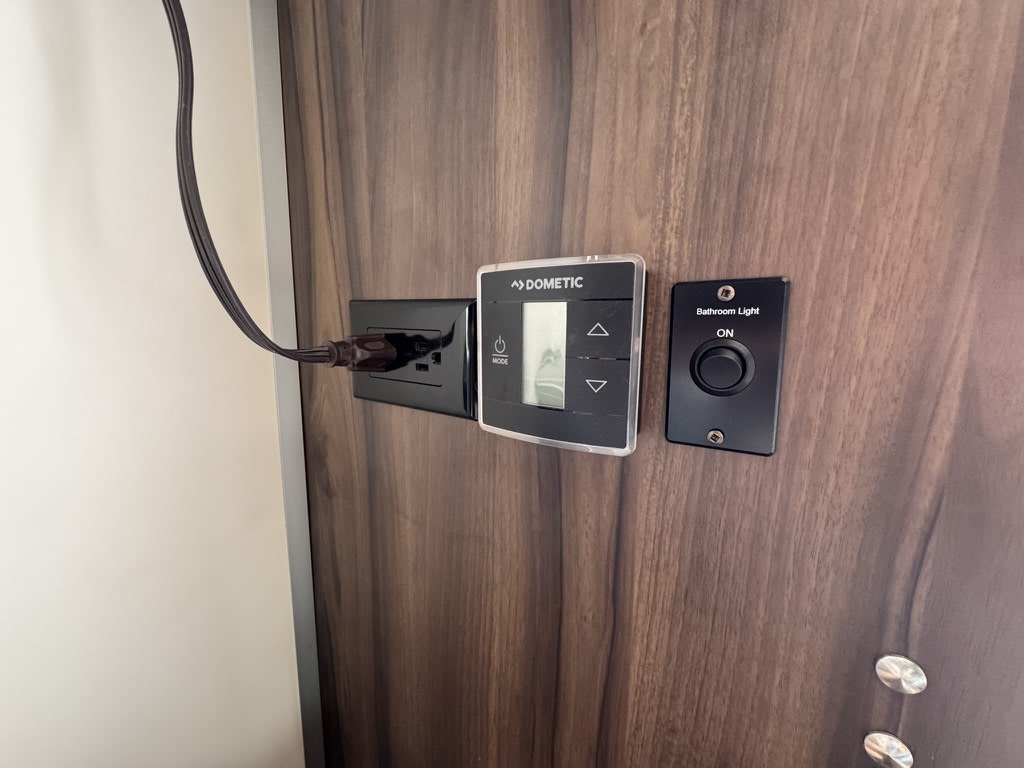 RV rental interior controls: Dometic thermostat, plugged-in outlet, and bathroom light switch on a wood panel.