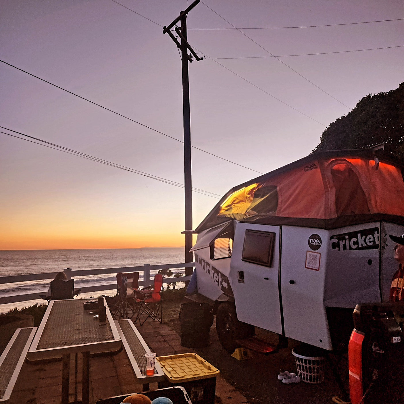 A unique Cricket **RV rental** with its pop-up tent glows at sunset by the ocean, with people enjoying beachside camping.