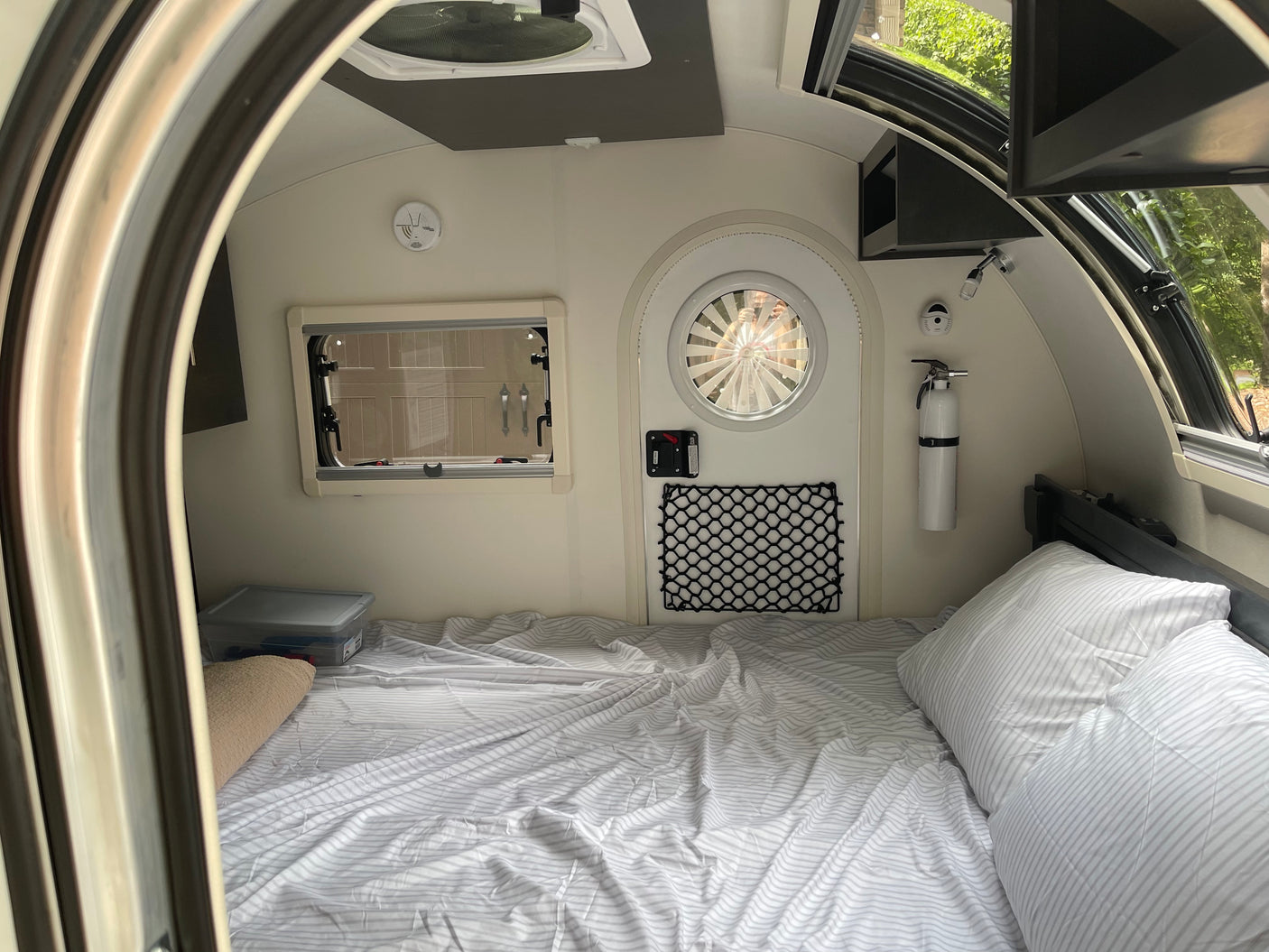 RV rental bedroom view: a compact space with a striped bed, side window, round door, fire extinguisher & ceiling vent.