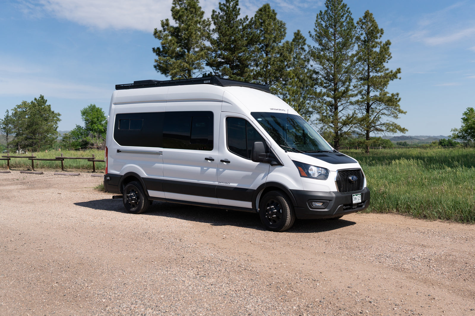 White adventure camper van, ideal for RV rental and outdoor road trips, parked on a gravel path with trees and blue sky.