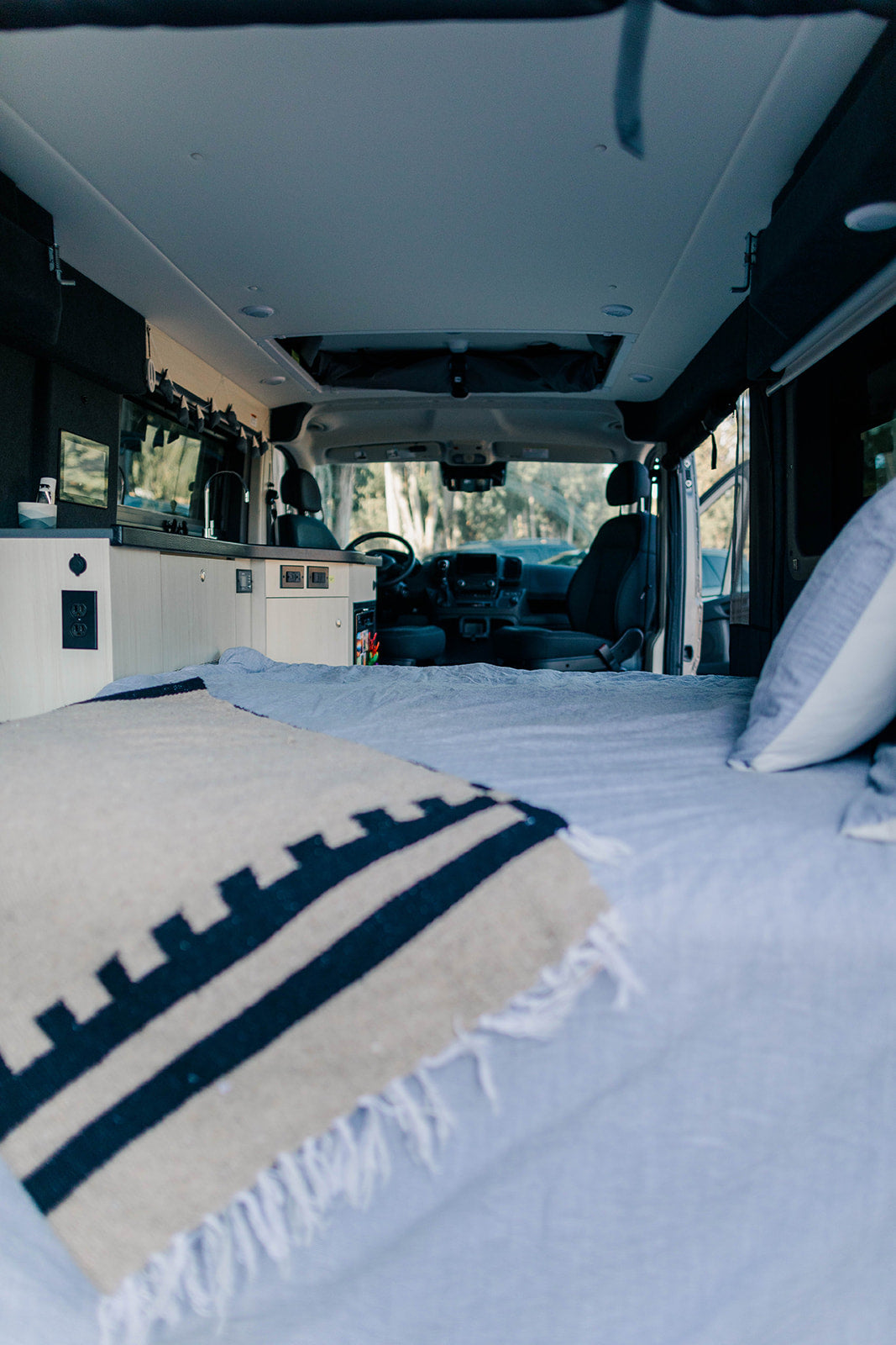 Inside a cozy RV rental: a comfortable bed with a patterned blanket leads to a built-in kitchenette and the driver's area beyond.