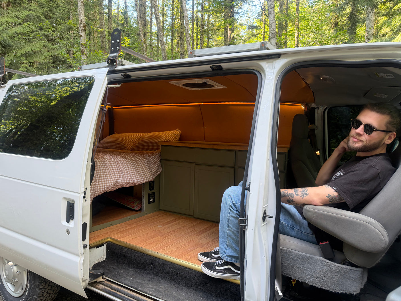 A man sits in his converted RV rental van, showcasing its custom bed, cabinets, and lighting for outdoor adventures.