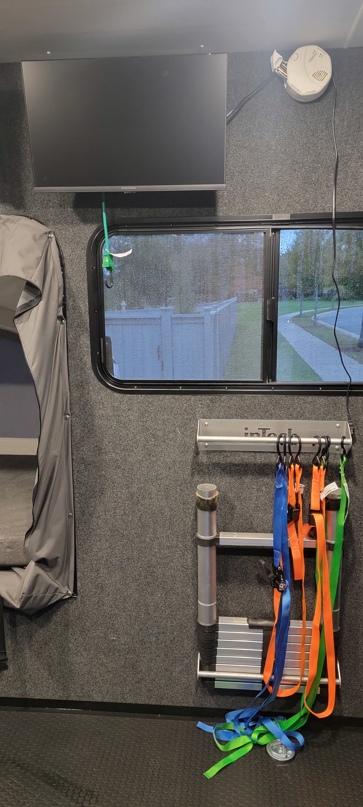 RV rental interior view: Mounted TV, rainy window, utility ladder, and tie-down straps stored on carpeted walls.