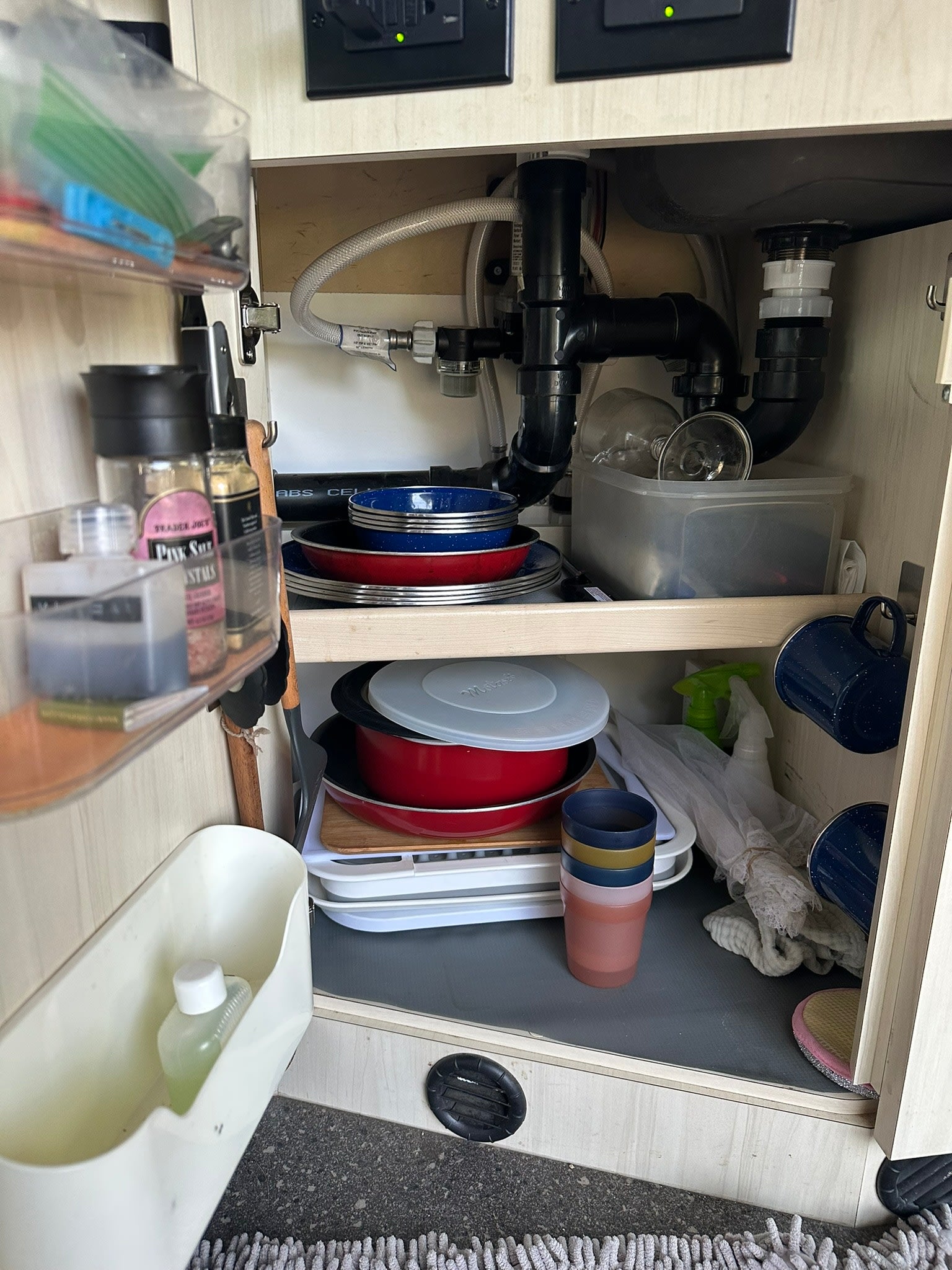 Inside an RV rental, this under-sink cabinet reveals compact kitchen storage with plumbing, dishes, pots, spices, and cleaning supplies.