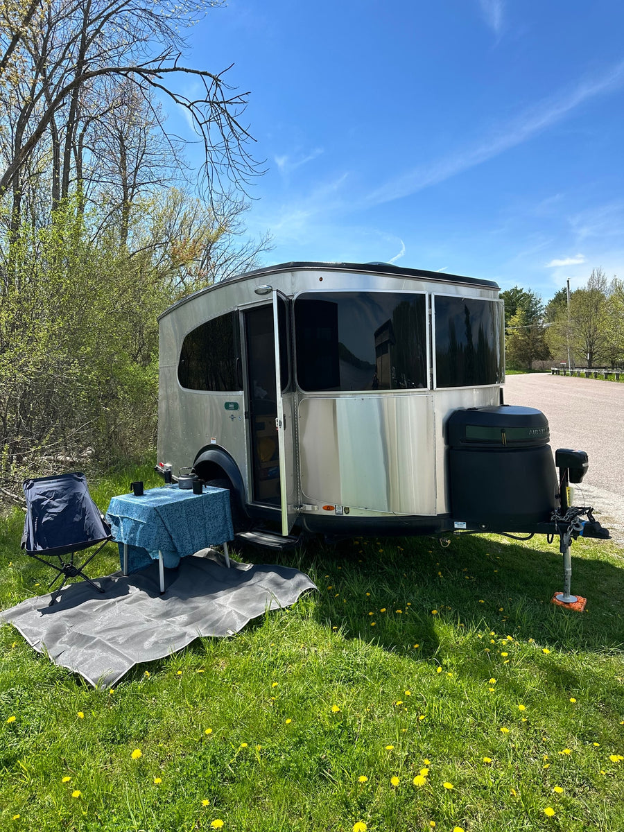 Perfect day for an rv rental! A silver travel trailer, table, & chair sit on a sunny, dandelion-filled lawn.