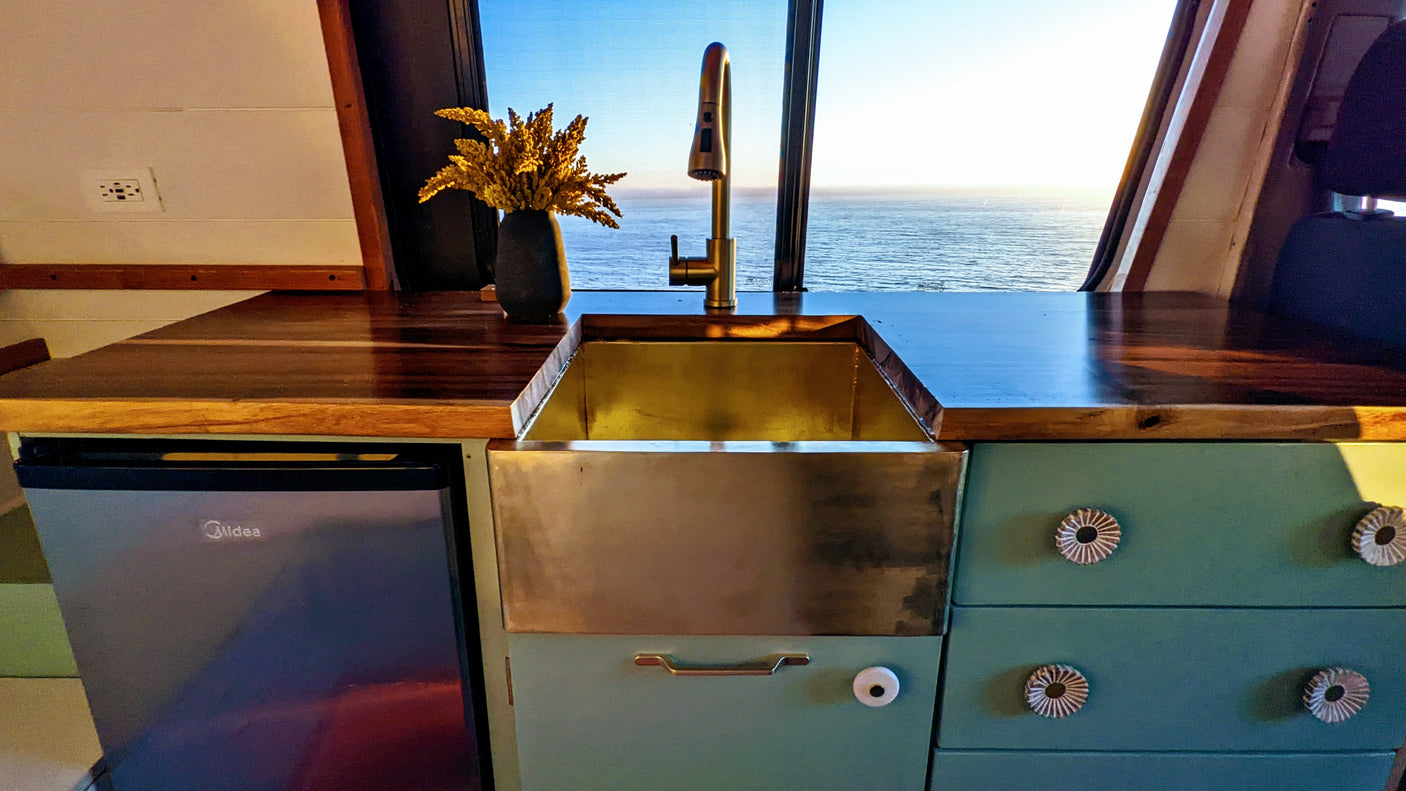 RV rental kitchen: wood counters, gold faucet, teal cabinets, and an ocean view through the large window. Features a Midea fridge.