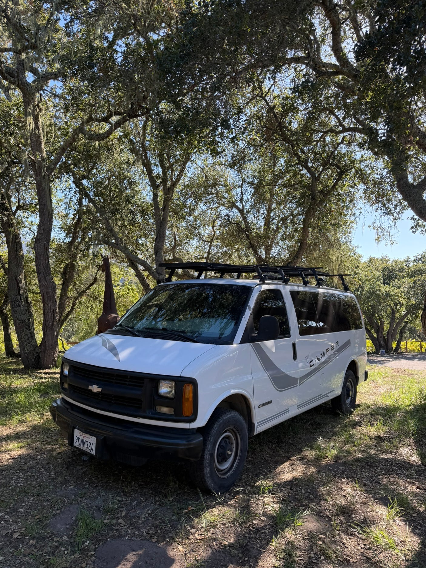White Chevy camper van, great for an rv rental, parked under mossy oak trees with a giraffe statue in background.