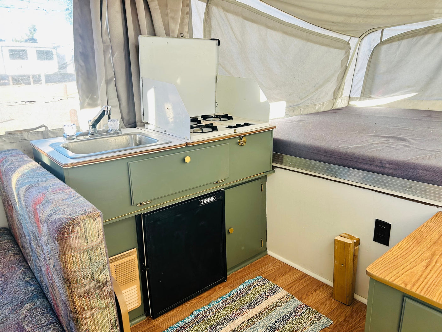 RV rental pop-up camper interior: sage green kitchenette with sink, stove, fridge, upholstered seating, and a bed.