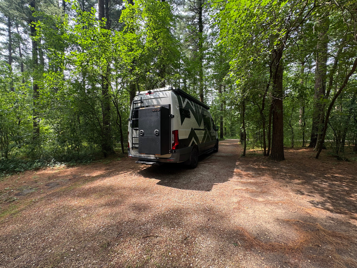 White camper van RV rental parked on a gravel path in a sunny forest, ready for adventure.
