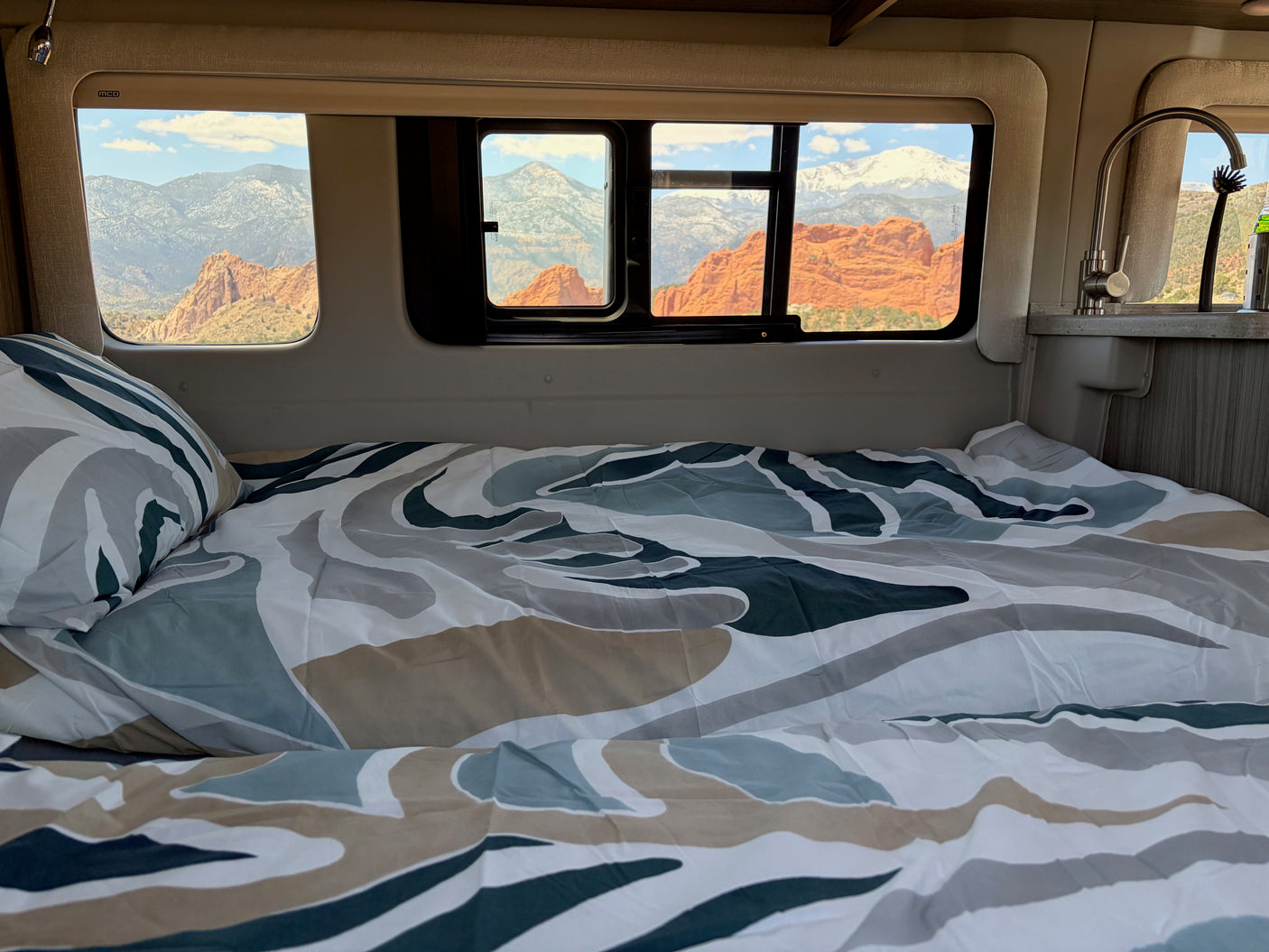 Enjoy breathtaking red rock and snow-capped mountain views from a cozy RV rental bed with patterned bedding and nearby sink.