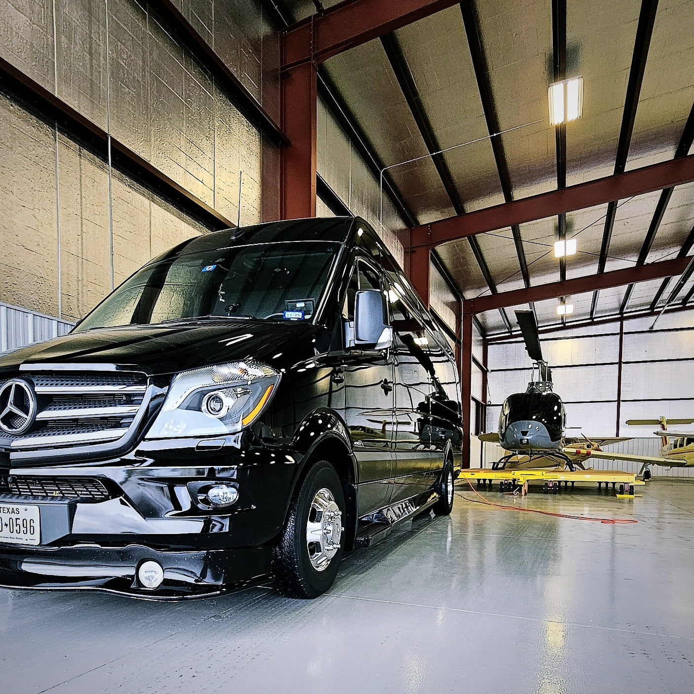 This sleek Mercedes Sprinter RV rental van stands ready in a hangar, sharing space with a helicopter & aircraft.