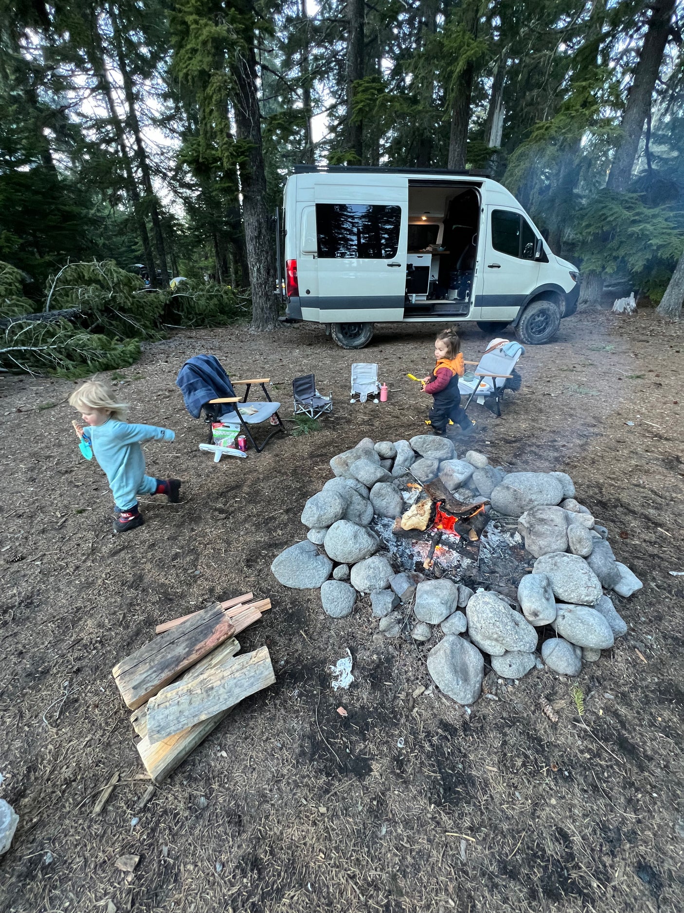 An RV rental is parked at a wooded campsite, where two young children play near a large, stone-ringed campfire.