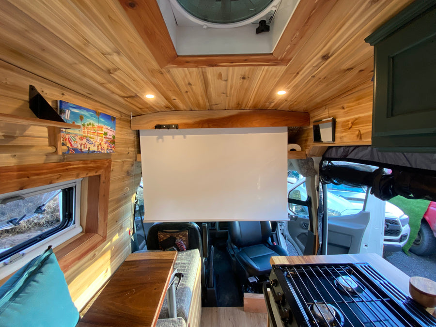 Cozy camper van interior for RV rental, featuring wood accents, a projector screen, and compact kitchen.