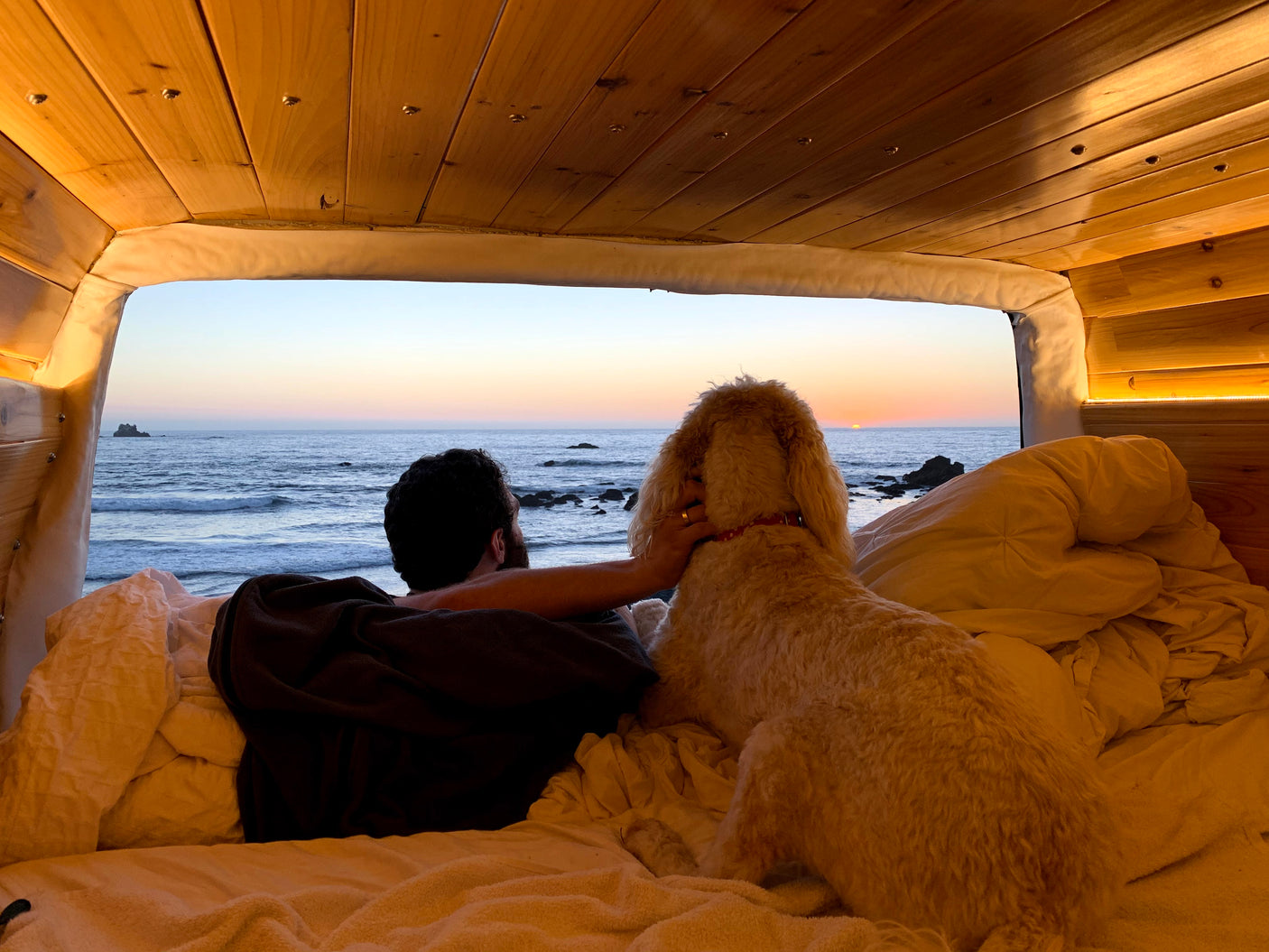 From a comfortable RV rental bed, a man and his dog gaze at a serene ocean sunset.