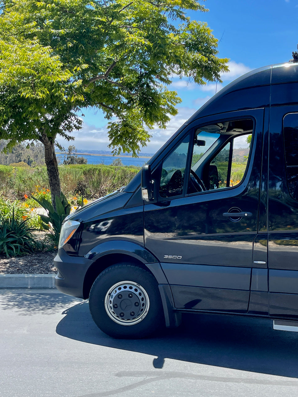 A black Mercedes Sprinter van, ready for your next RV rental trip, parked near a tree overlooking the ocean.