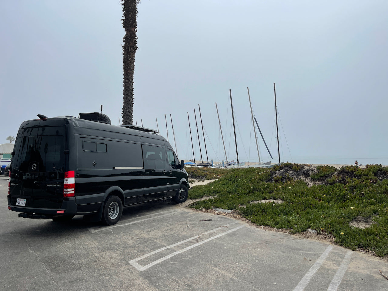 A sleek black Mercedes Sprinter RV rental parked on a paved lot by a sandy beach with sailboats and a misty sky.