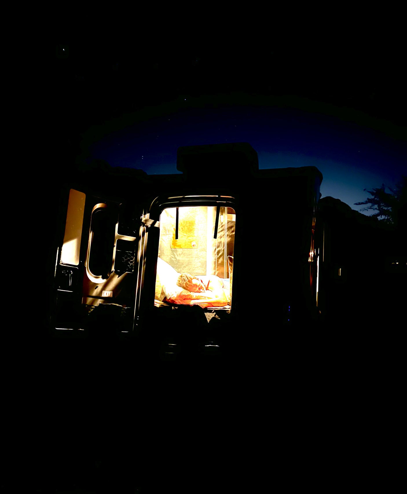 An RV rental at night, showing a person sleeping inside a brightly lit bed area. Stars dot the dark sky above the camping scene.